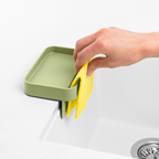 Blim+ Battigia Sponge Holder in green on a sink edge with a hand holding a yellow sponge