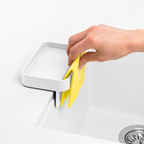 Blim+ Battigia Sponge Holder in white on a sink edge with a hand holding a yellow sponge