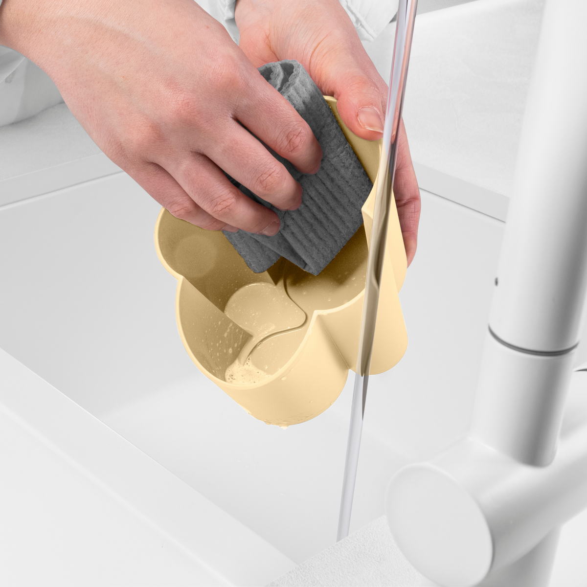 Blim+ Gocciolo Cutlery Drainer in pale yellow, with a hand holding a grey sponge beside a tap