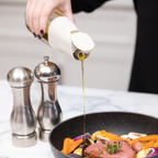 Zuutii Oil Dispenser in cream, pouring oil into a pan beside salt and pepper mills