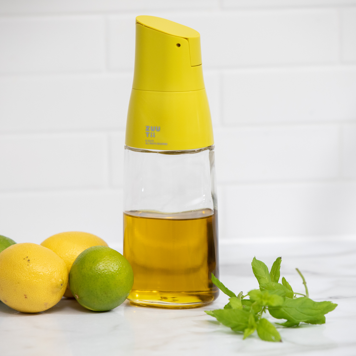 Zuutii Oil Dispenser in yellow with olive oil beside lemons, a lime and mint
