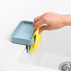 Blim+ Battigia Sponge Holder in blue on a sink edge with a hand holding a yellow sponge
