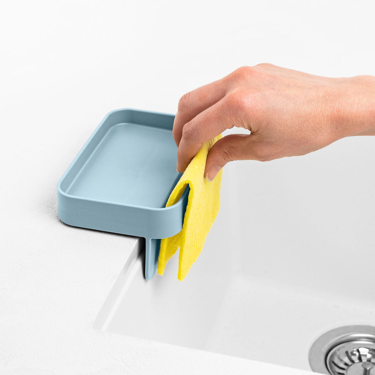 Blim+ Battigia Sponge Holder in blue on a sink edge with a hand holding a yellow sponge