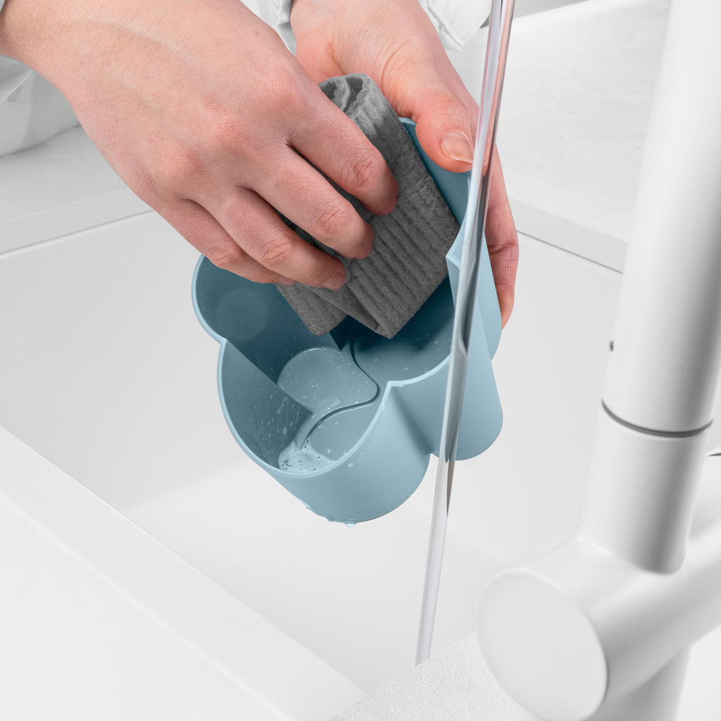 Blim+ Gocciolo Cutlery Drainer in blue, with a hand holding a grey sponge beside a tap
